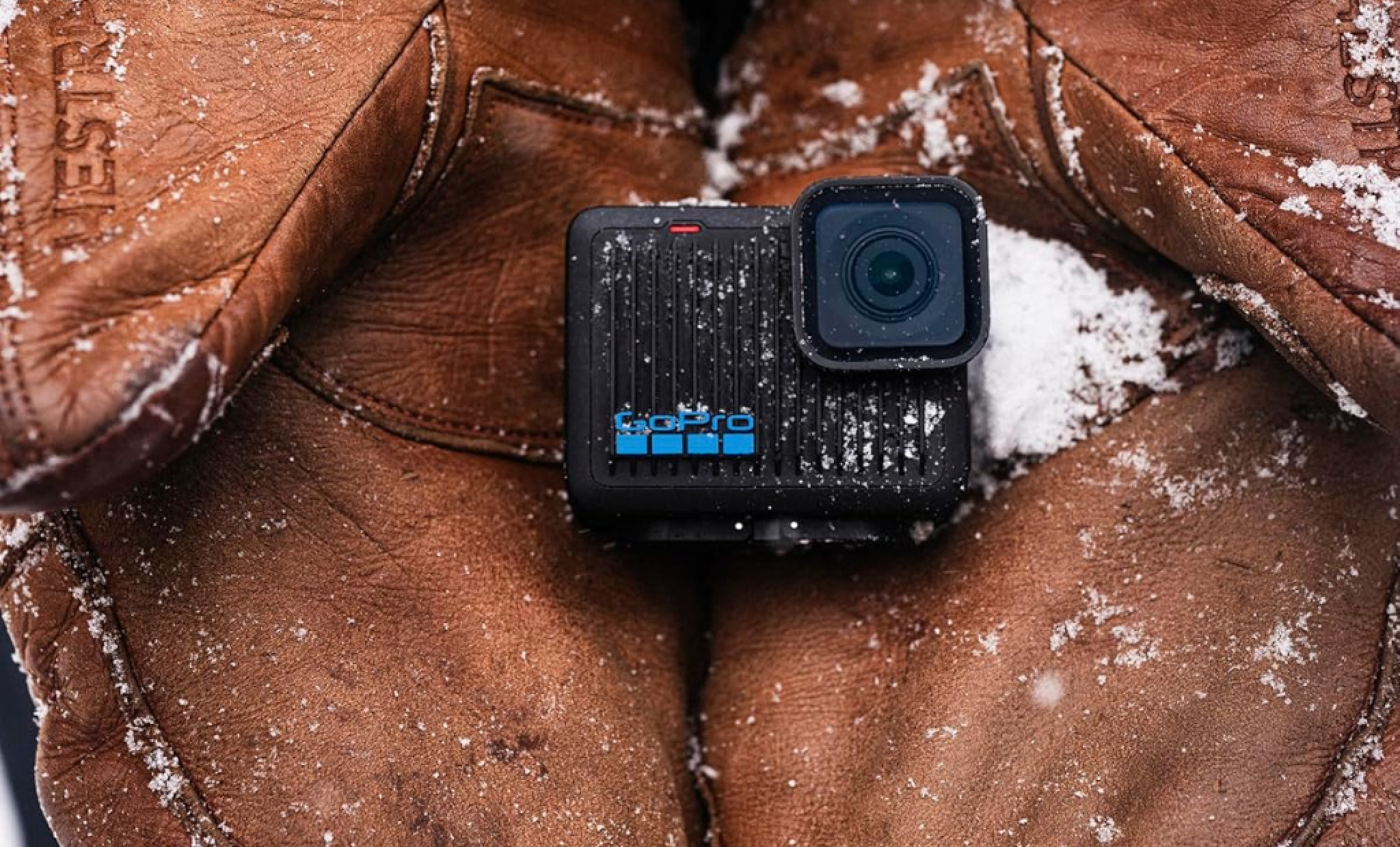Uma pessoa com roupa e luvas de neve, na neve, segunda com as duas palmas da mão juntas, uma câmera de ação go pro que está envolta em neve.